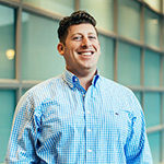 Headshot of a smiling person wearing a blue checkered shirt, standing indoors with a glass wall in the background.