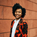 A person with curly hair smiles while wearing a vibrant floral-patterned jacket over a white shirt, standing in front of a textured brick wall.