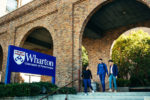 Three people walking by a sign for the Wharton School, University of Pennsylvania, in front of a brick building with arches.