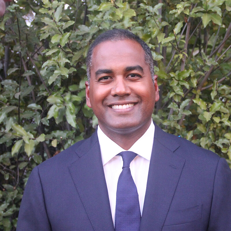 A headshot of a person wearing a navy suit and tie with a white shirt, smiling, with a background of green leaves.