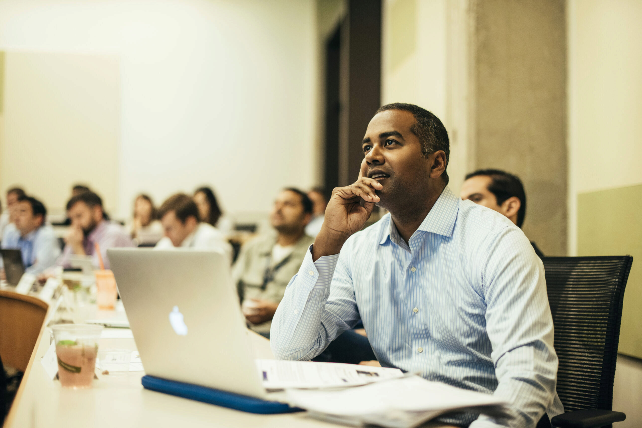 A person in a classroom setting attentively listening, with a laptop and papers in front, surrounded by others in a similar environment.