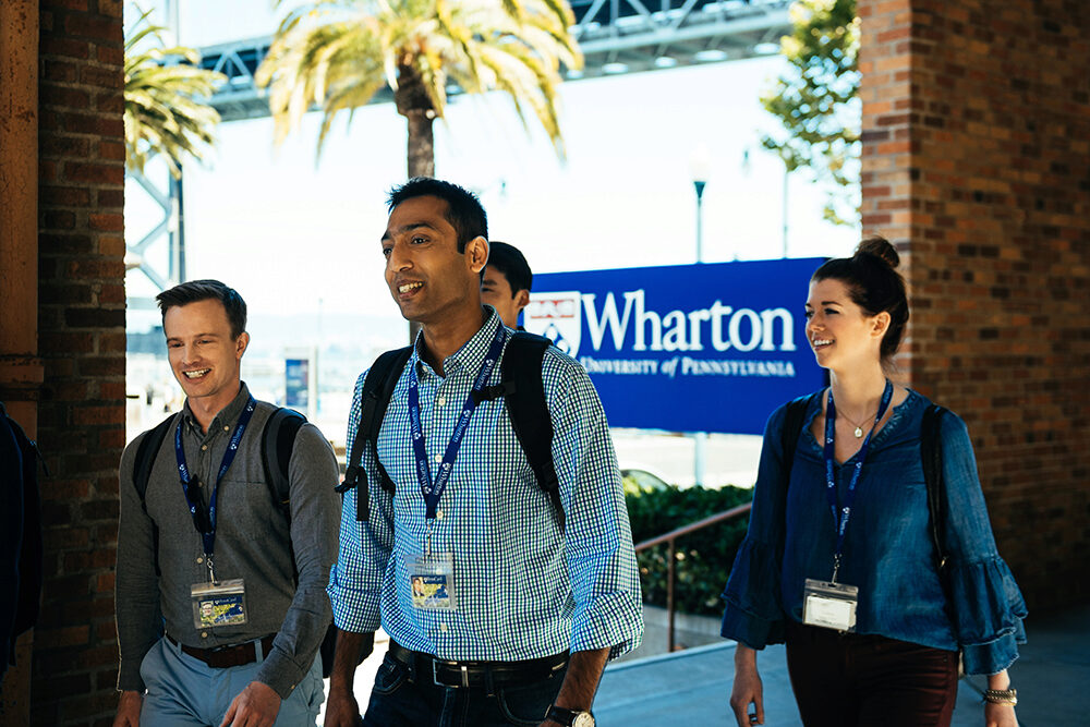 A group of people walking outside, wearing lanyards with ID badges. A Wharton School of the University of Pennsylvania sign is visible behind them, with palm trees and part of a steel structure in the background.