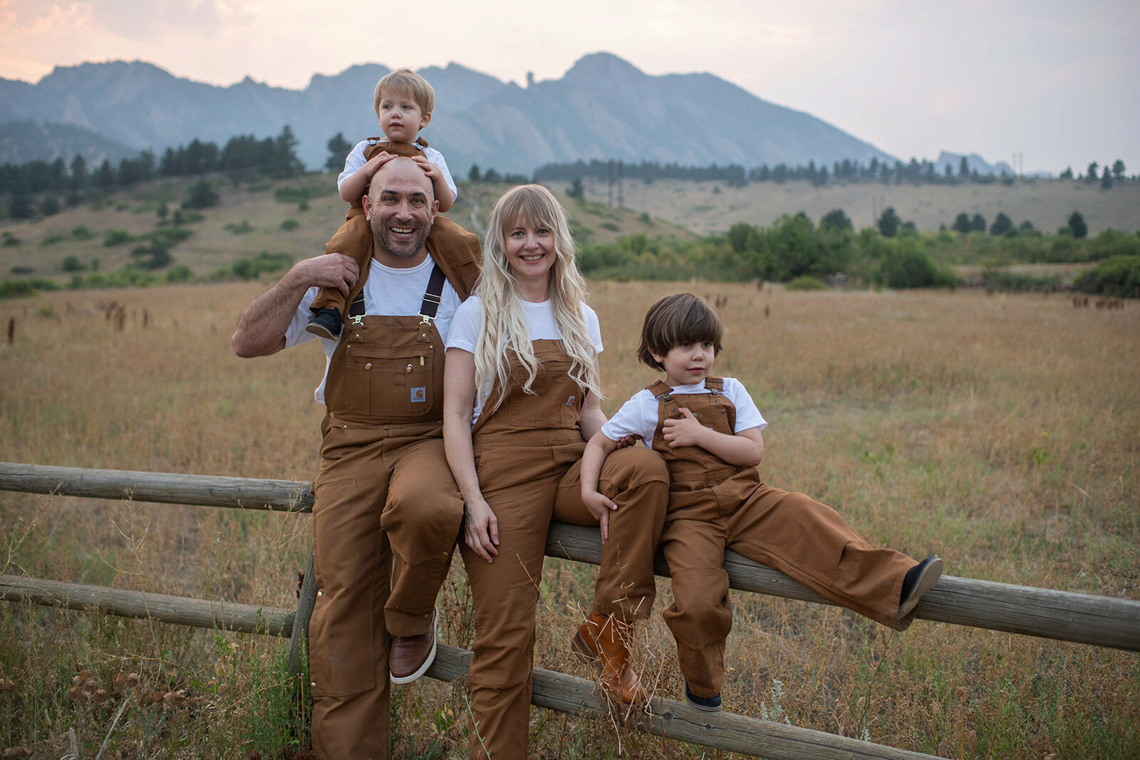 A family of four wearing brown overalls and white shirts is sitting on a wooden fence in a grassy field with mountains in the background. One adult holds a child on their shoulders.