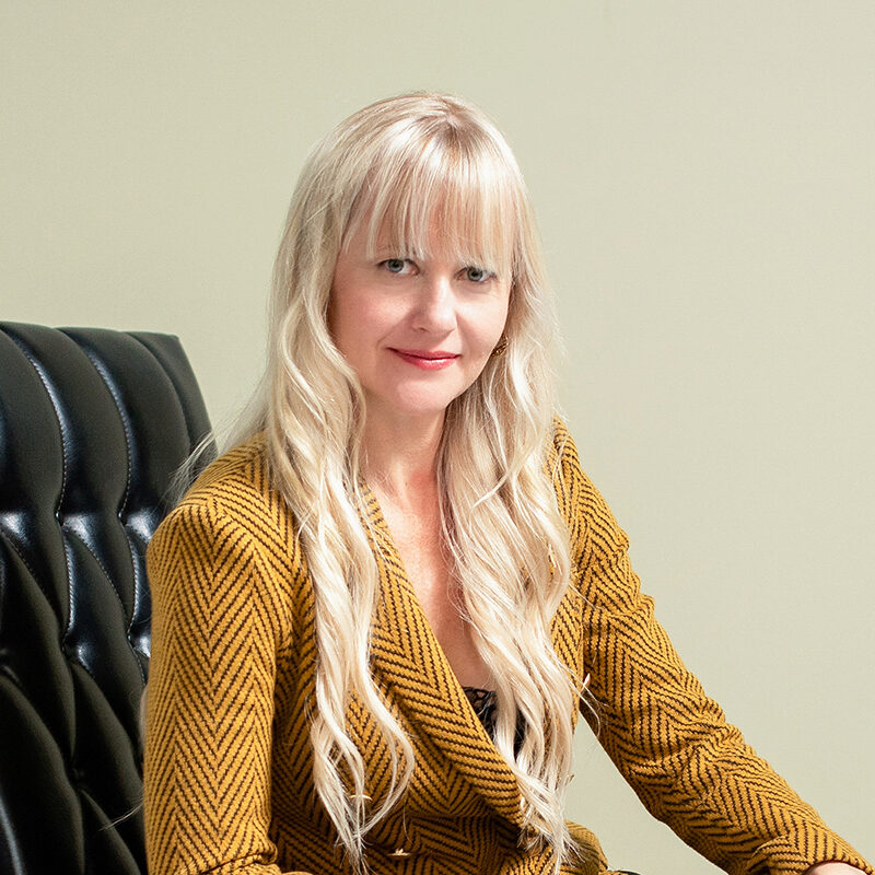 A person with long blond hair, wearing a mustard-colored striped blazer, sitting on a black leather chair against a light background.