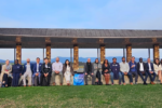 A group of people posed in front of a stone structure with a lawn in the foreground. They are seated and standing, dressed in casual to business attire, in an outdoor setting.